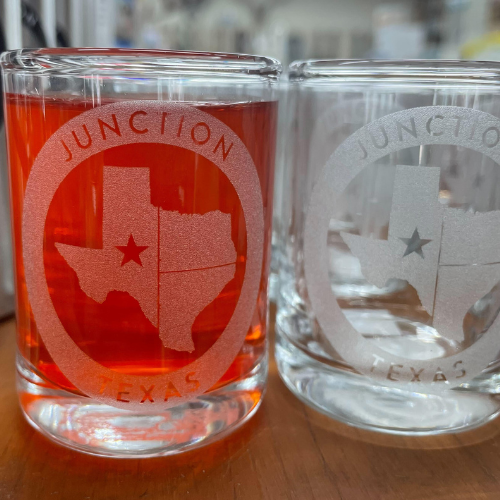 Two Junction Texas branded shot glasses: one filled with red liquid and the other empty. Both glasses feature a white design with a circle around the state of Texas, including a star on Junction and the text 'Junction Texas'.