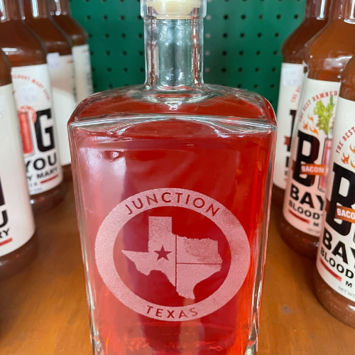 Junction Texas branded glass bottle with a cork, containing red liquid. The design features a white circle with the state of Texas and a star on Junction, with 'Junction Texas' written in white around the state.